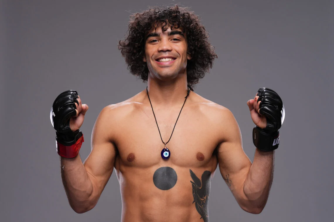 MMA fighter smiling with raised fists, wearing fingerless gloves, a blue eye pendant and torso tattoos against a gray studio background