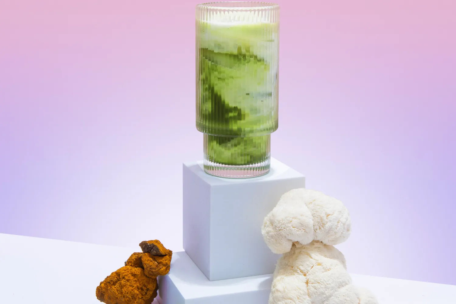 Tall ribbed glass of mushroom matcha on a white pedestal with lion’s mane and chaga mushrooms against a pink to purple gradient background