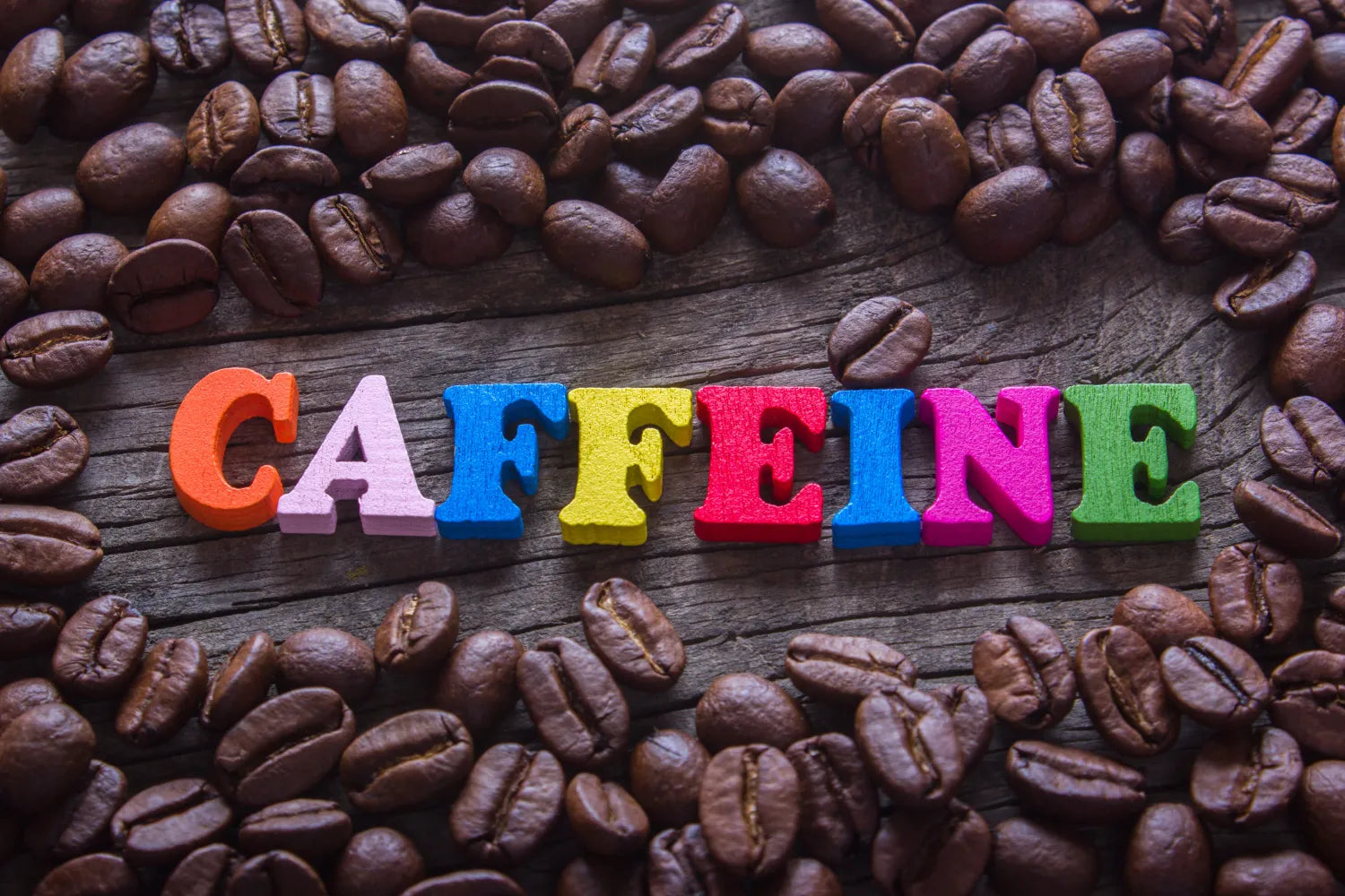 Colorful letters spelling CAFFEINE surrounded by roasted coffee beans on a weathered wooden surface