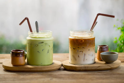 Two iced drinks, a green matcha latte and layered iced coffee, served in glass jars with brown straws on wooden serving boards