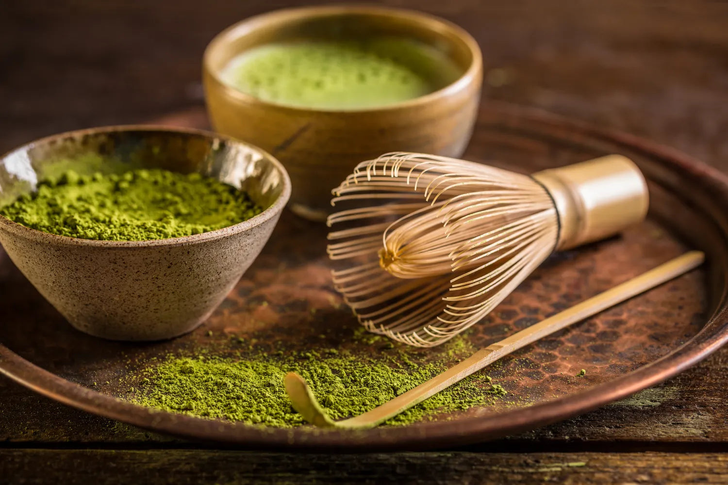 Bowls of bright green matcha powder and whisked matcha with a bamboo whisk and scoop on a rustic tray