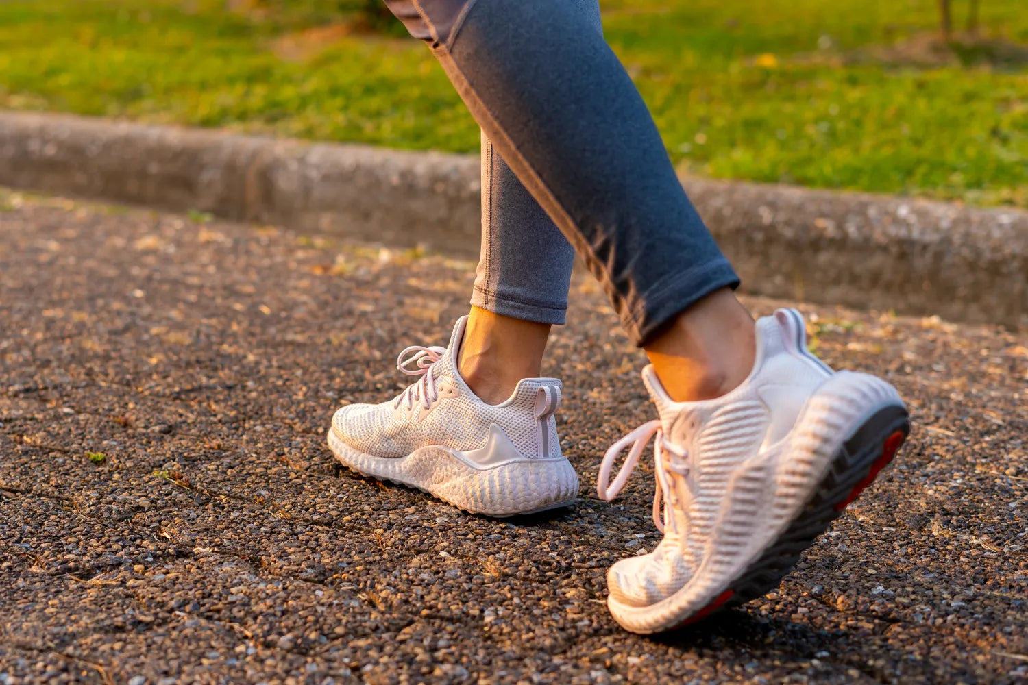 Person walking on a paved path wearing white running shoes and gray leggings at sunset in a park