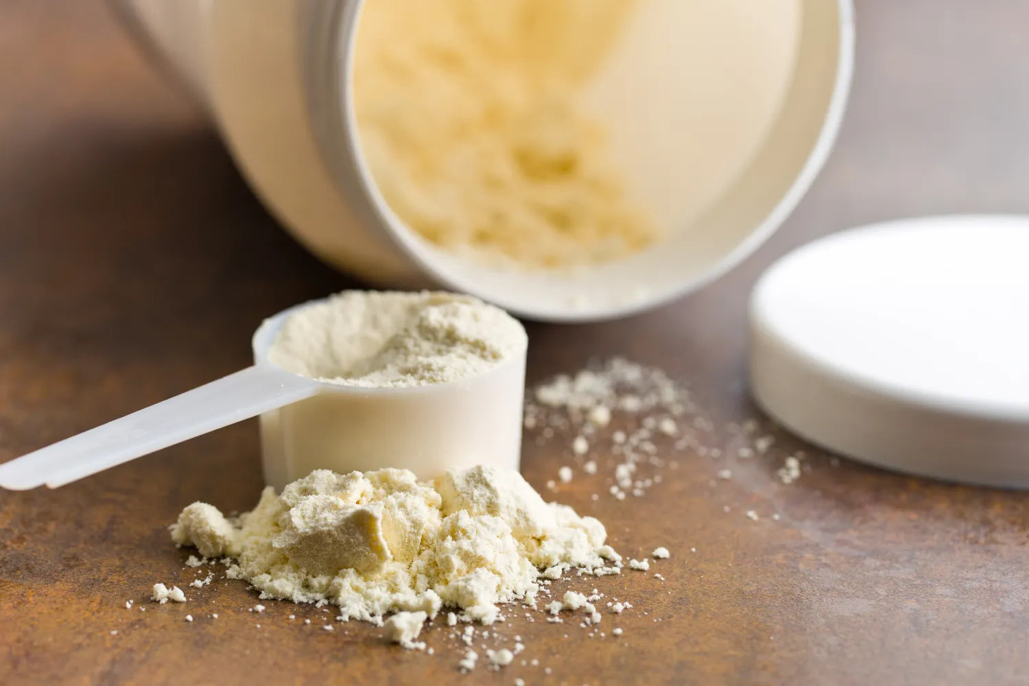 Close-up of a white scoop of protein powder next to an open white container with powder spilled on a brown tabletop