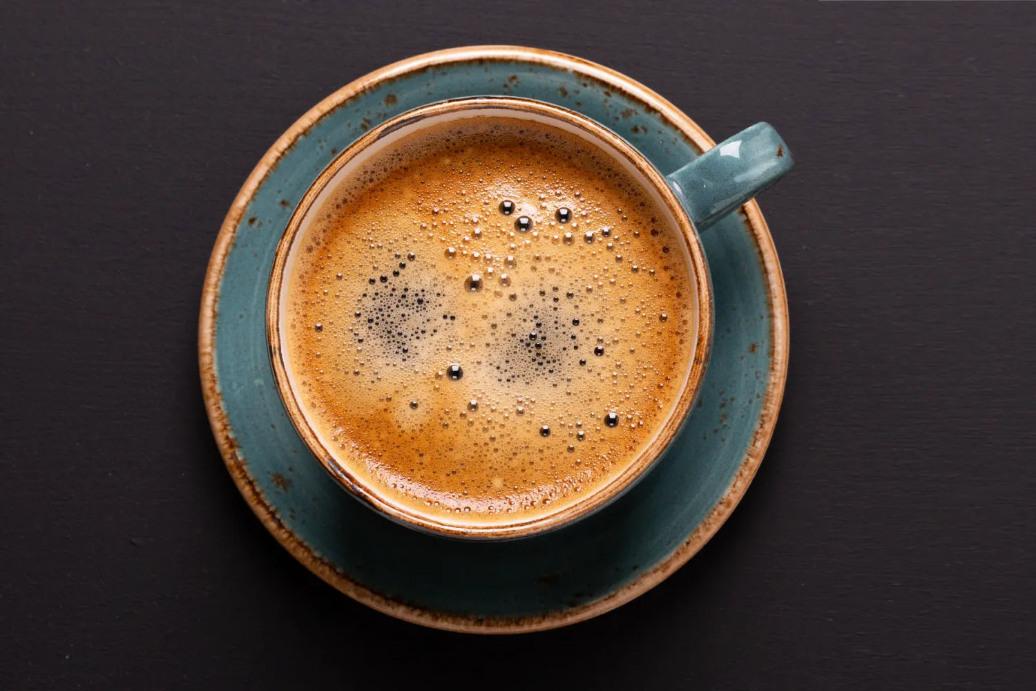 Top view of a cup of coffee with golden crema and bubbles in a blue ceramic cup and saucer on a dark table