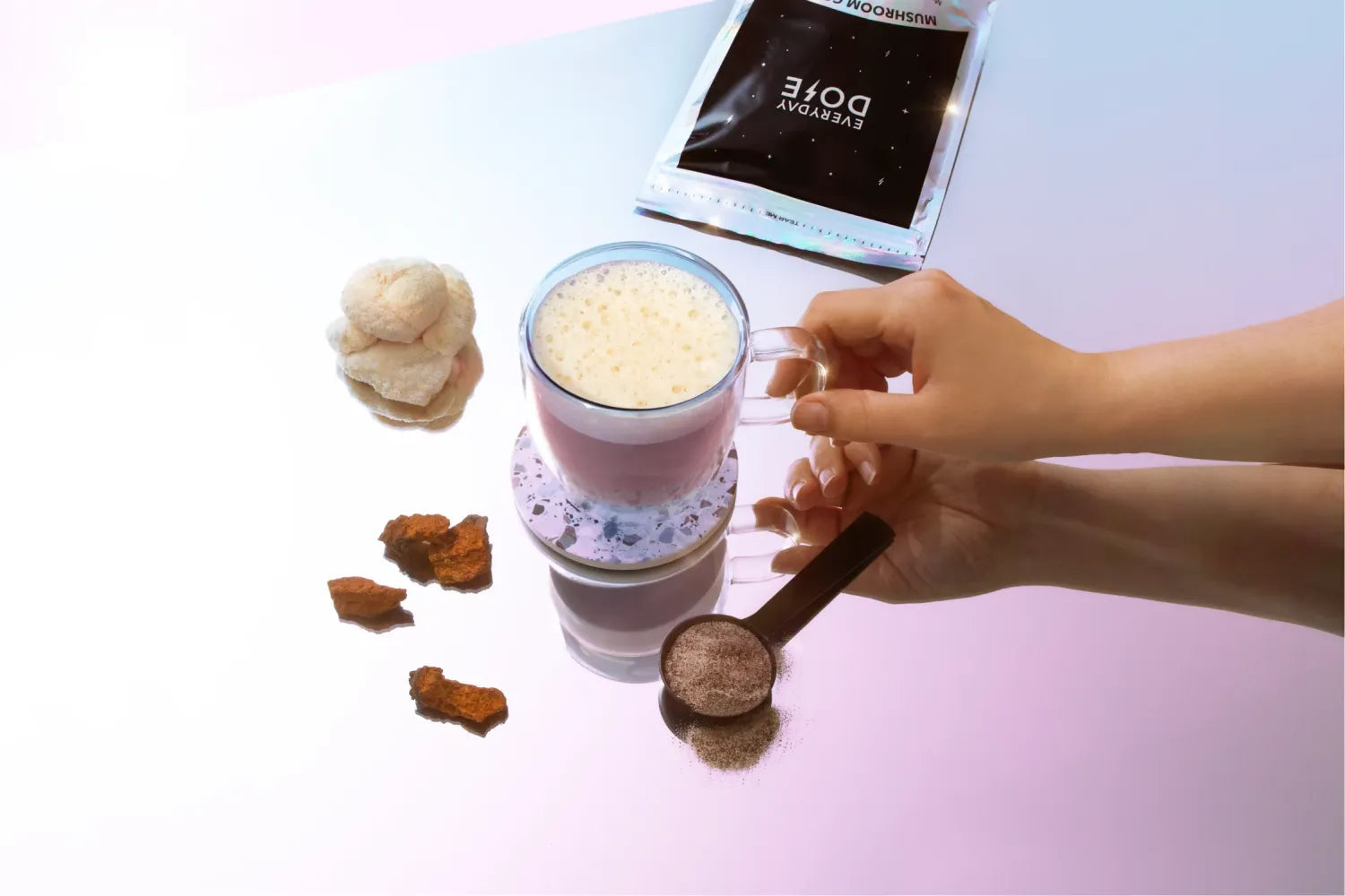 Top-down view of mushroom coffee latte in glass mug with froth, powder scoop, mushroom pieces and packet on pink surface