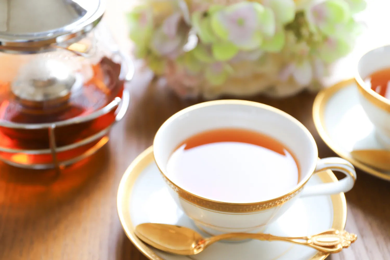 Tea cup on a saucer with gold trim beside a glass teapot and soft-focus flowers on a wooden table