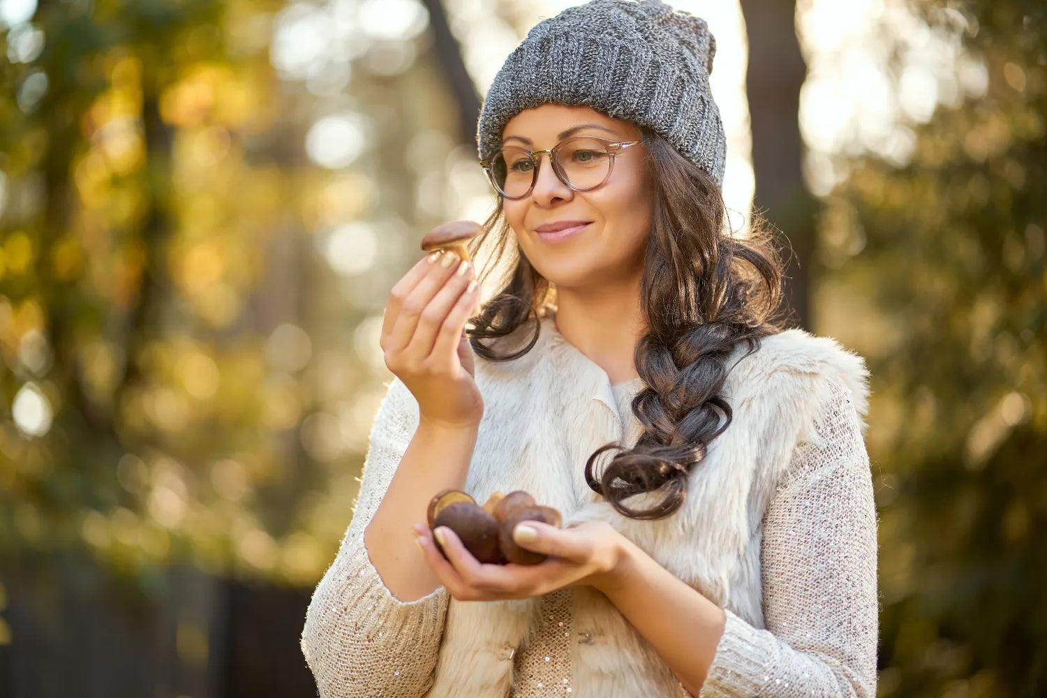 Woman outdoors holding and inspecting several brown mushrooms, wearing glasses, a knit hat, and a fur vest against soft autumn bokeh