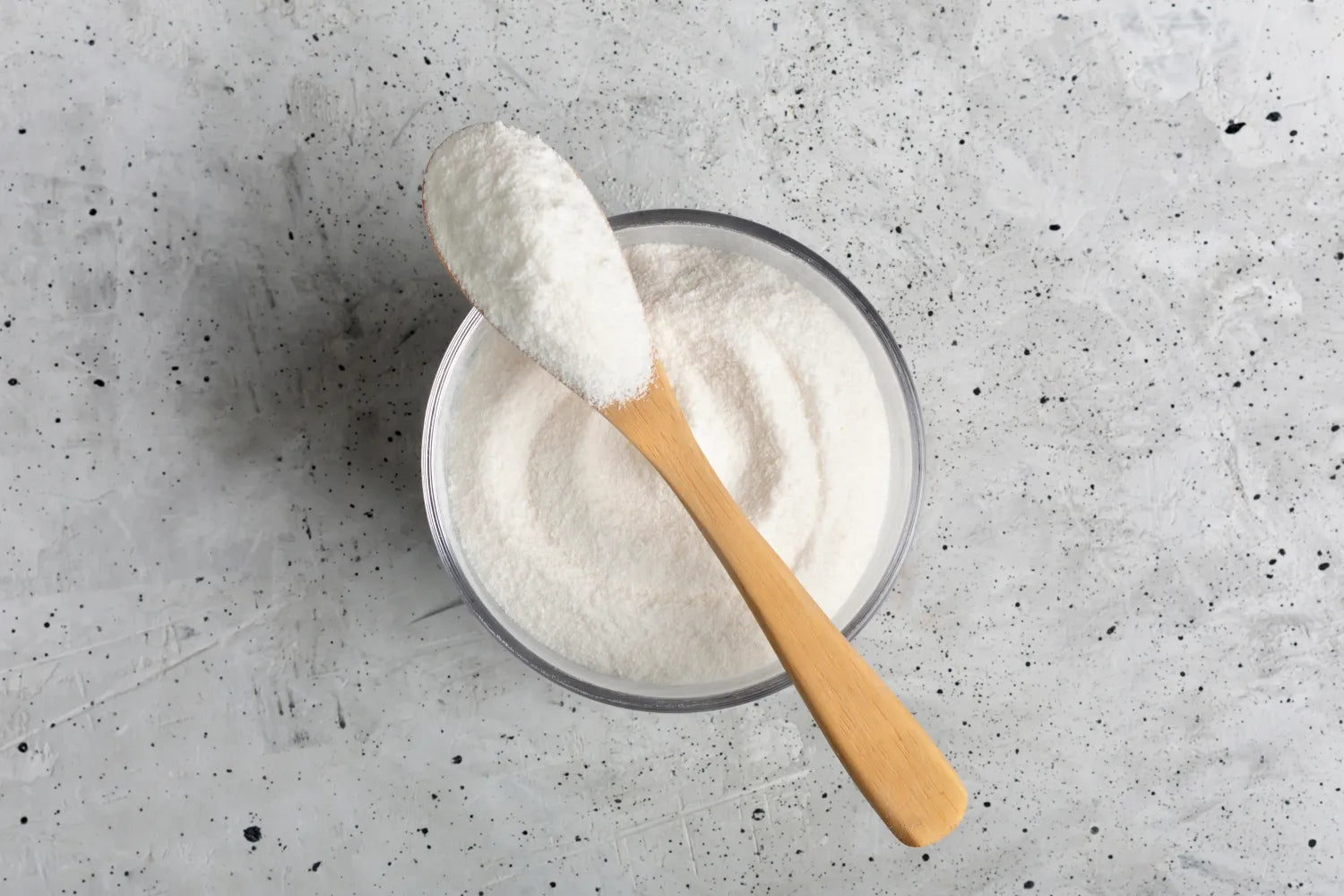 Wooden spoon scooping white powder from a glass bowl on a speckled gray surface