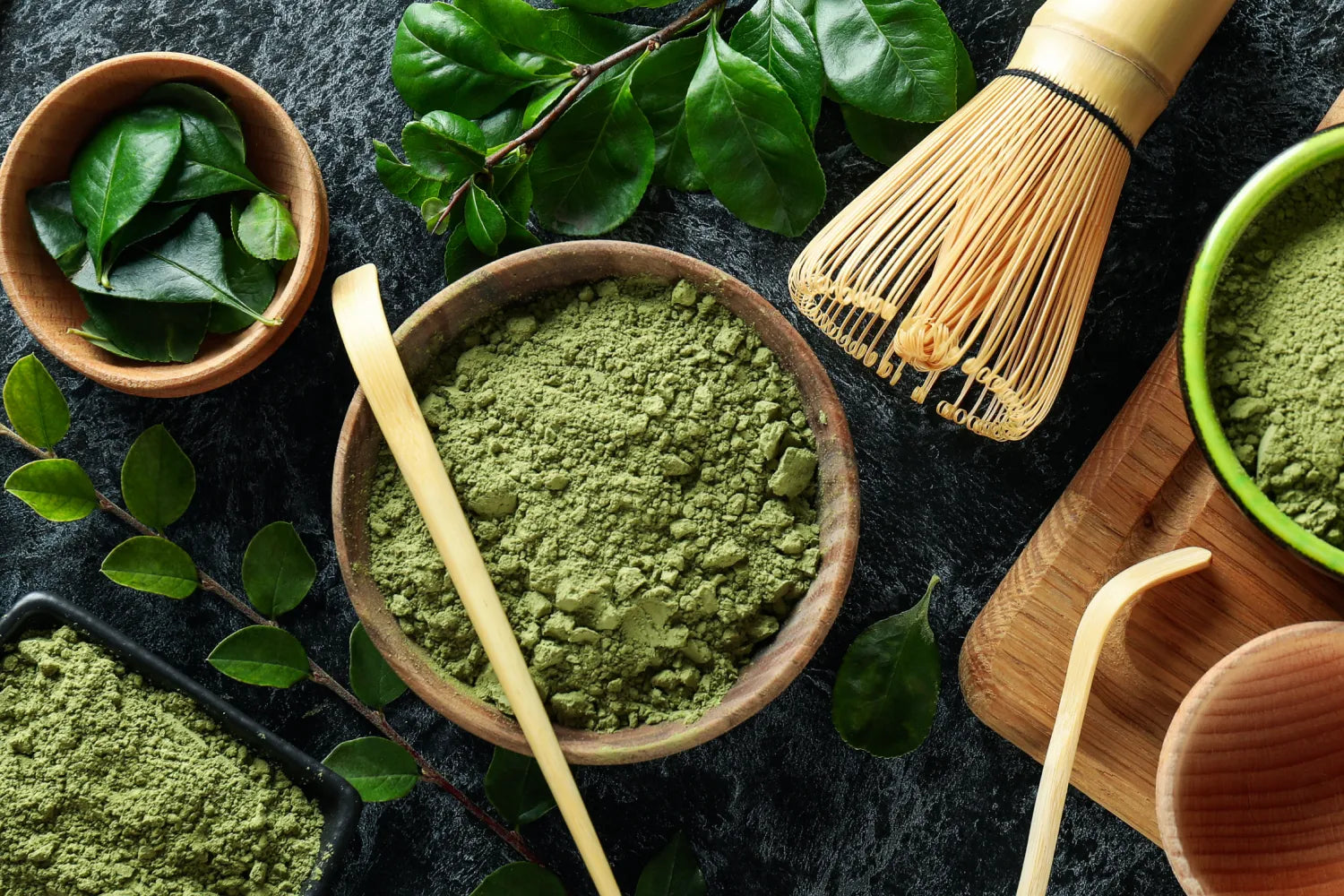 Overhead view of a bowl of bright green matcha powder with bamboo scoop and whisk on a dark stone surface