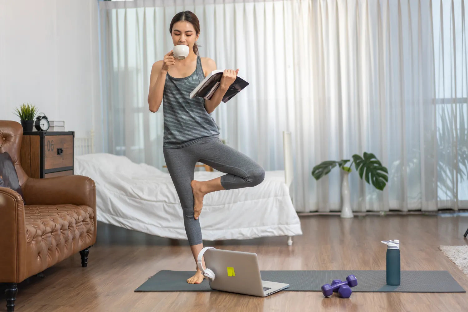 Woman balancing on one leg drinking coffee in a bedroom with yoga mat, laptop, dumbbells and water bottle