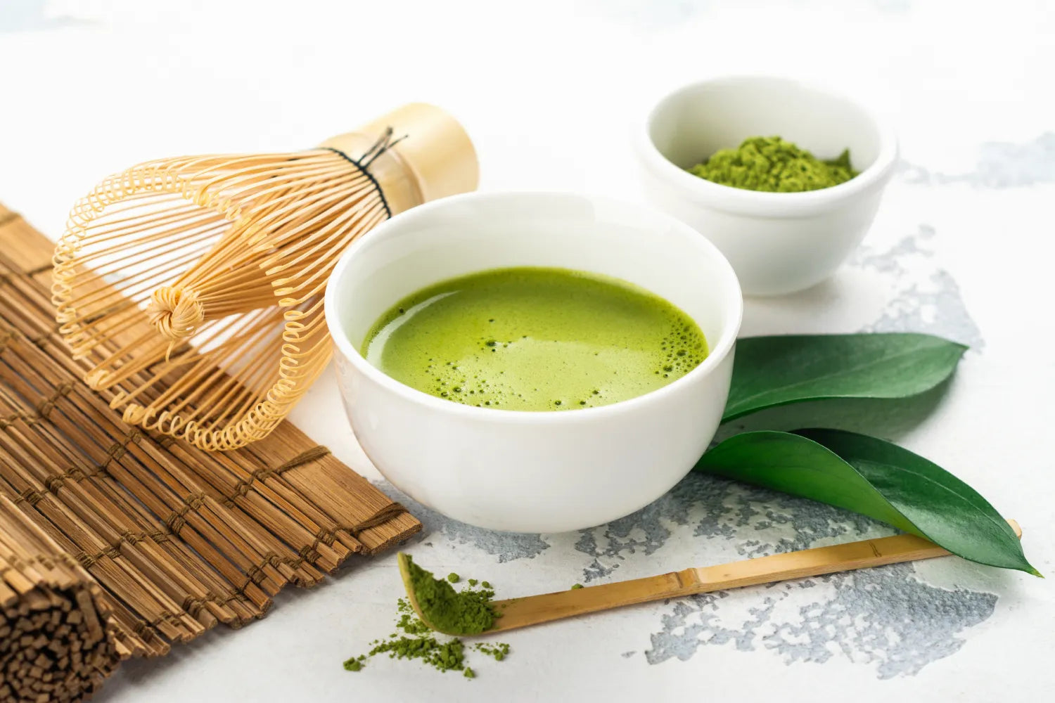 Bowl of bright green whisked matcha with bamboo whisk, scoop of powder, and tea leaves on a light surface