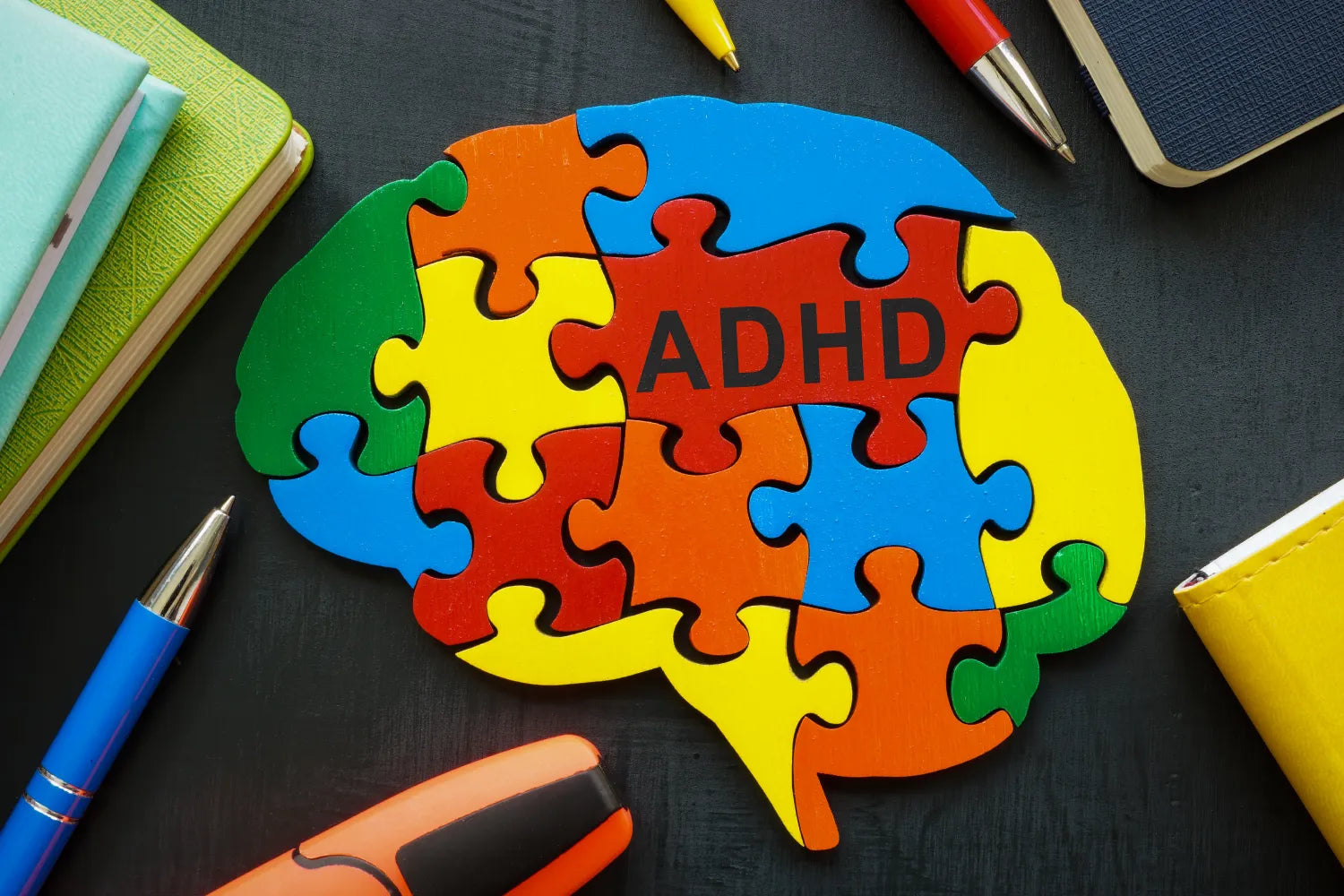 Illustration of a colorful jigsaw puzzle in the shape of a brain labeled ADHD, on a dark desk with pens and notebooks