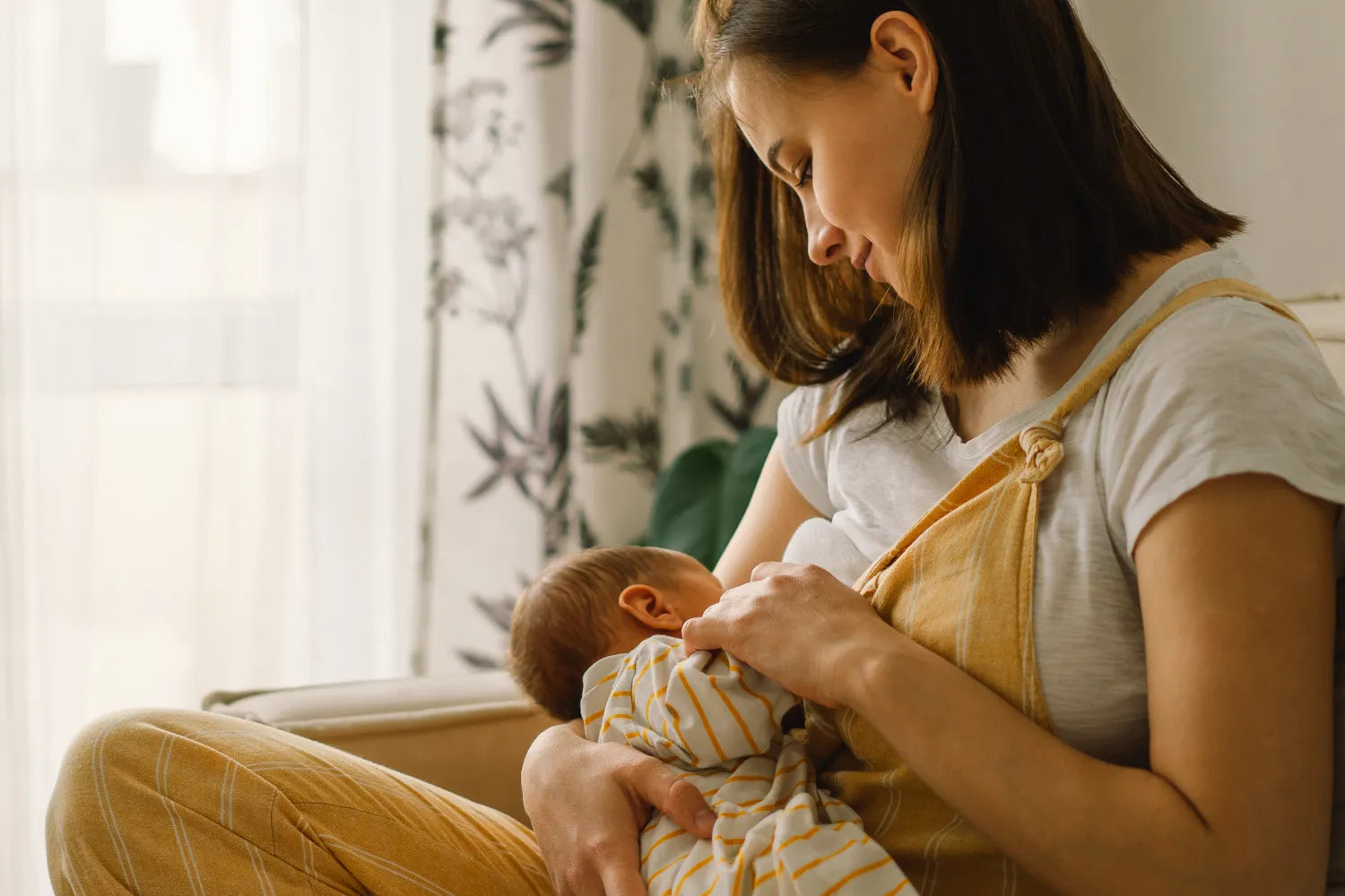 Mother breastfeeding a newborn seated in a sunlit room, wearing yellow overalls and cradling the baby in a white and yellow striped outfit