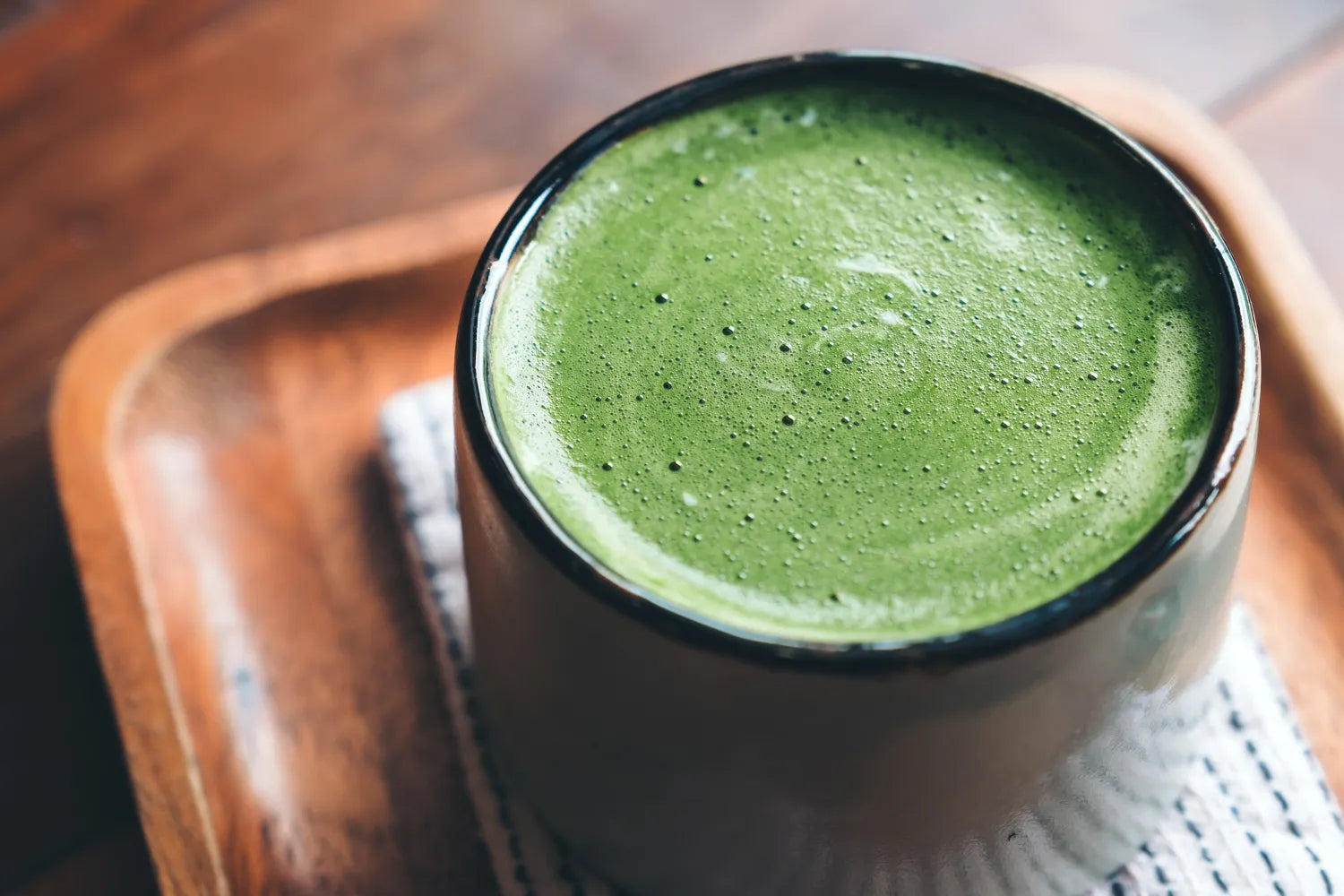 Top-down view of a frothy matcha latte in a ceramic cup showcasing bright green foam on a wooden tray