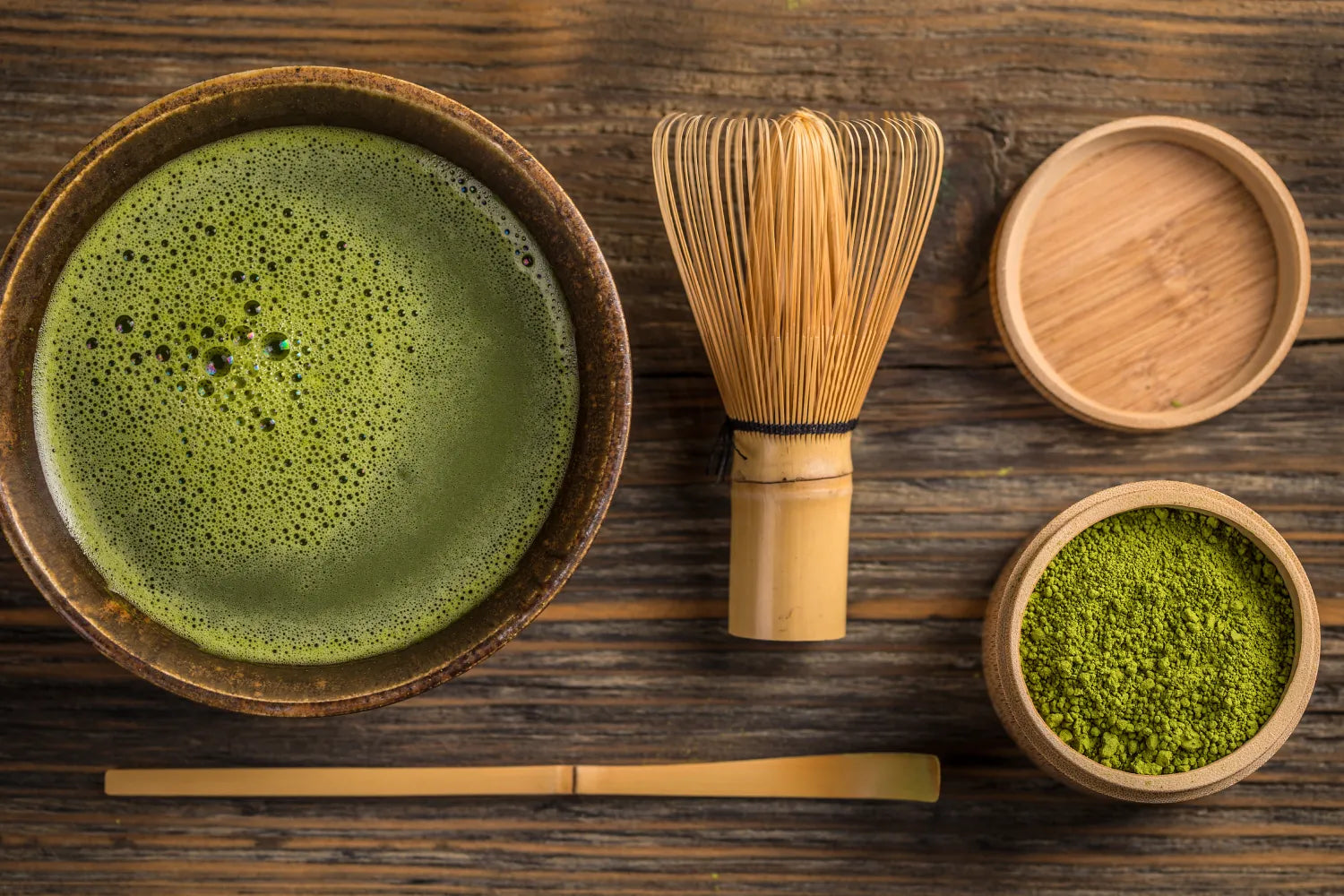 Top-down view of a frothy bowl of matcha next to a bamboo whisk, scoop, and container of matcha powder on a wooden table
