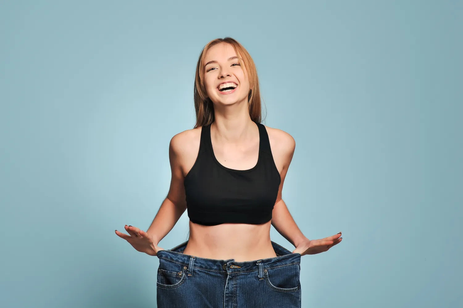 Young woman smiling in black sports bra holding oversized jeans to show weight loss against a light blue background