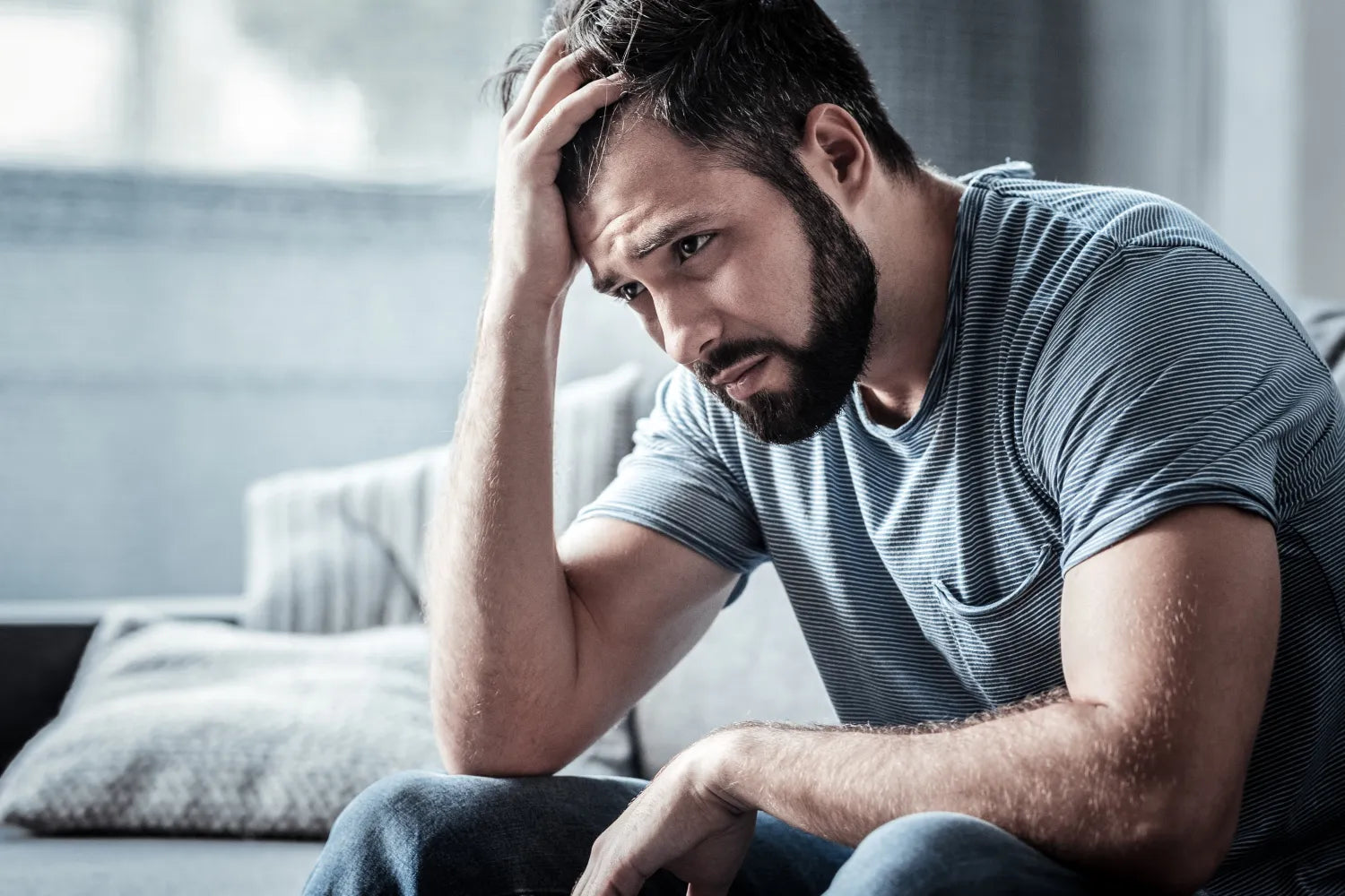 Bearded man sitting on a couch holding his head, looking distressed in a softly lit living room