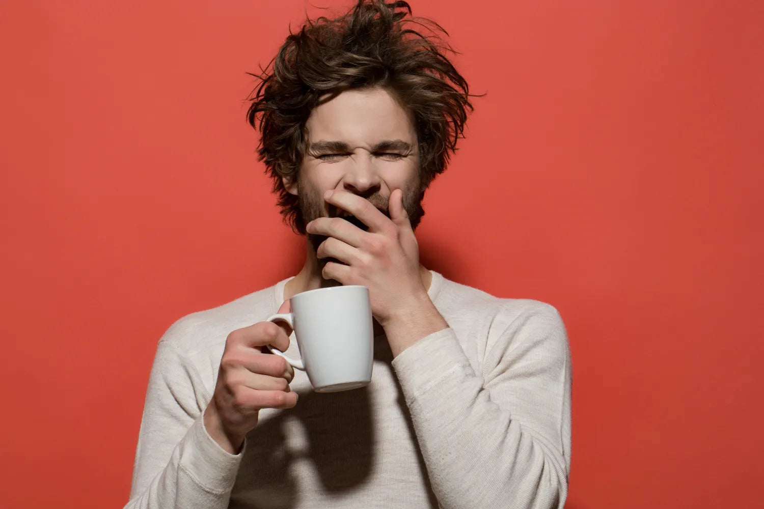 Man yawning while holding a white coffee mug against a red background