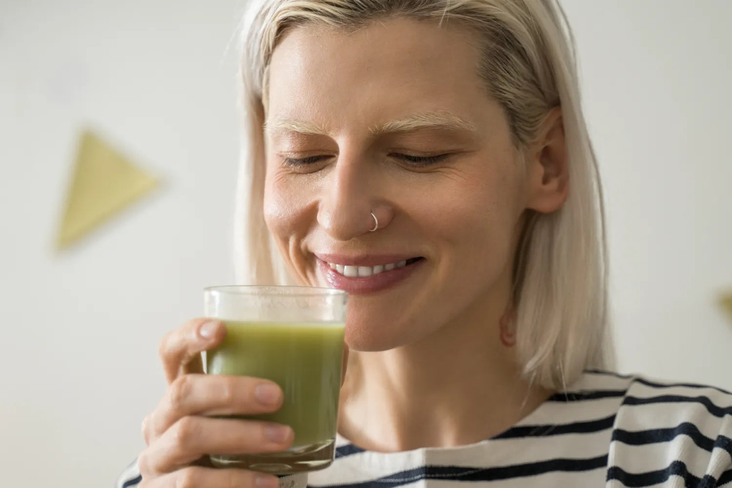 Close-up of a woman smiling as she holds and savors a glass of green matcha drink in a bright neutral kitchen