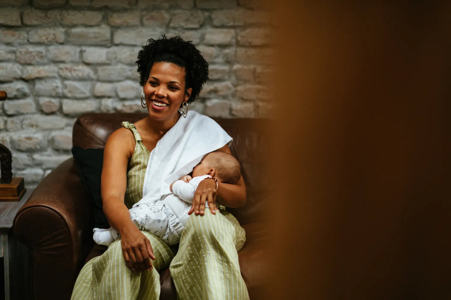 Smiling mother breastfeeding her infant on a leather couch with an exposed brick wall in a cozy living room