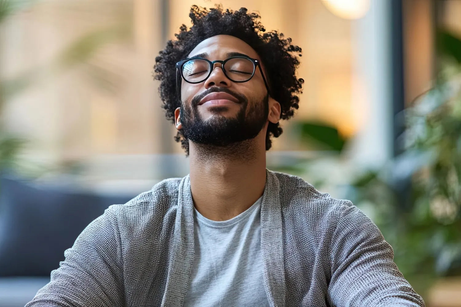Man with glasses sitting with eyes closed, taking a mindful breath in a softly lit living room with plants