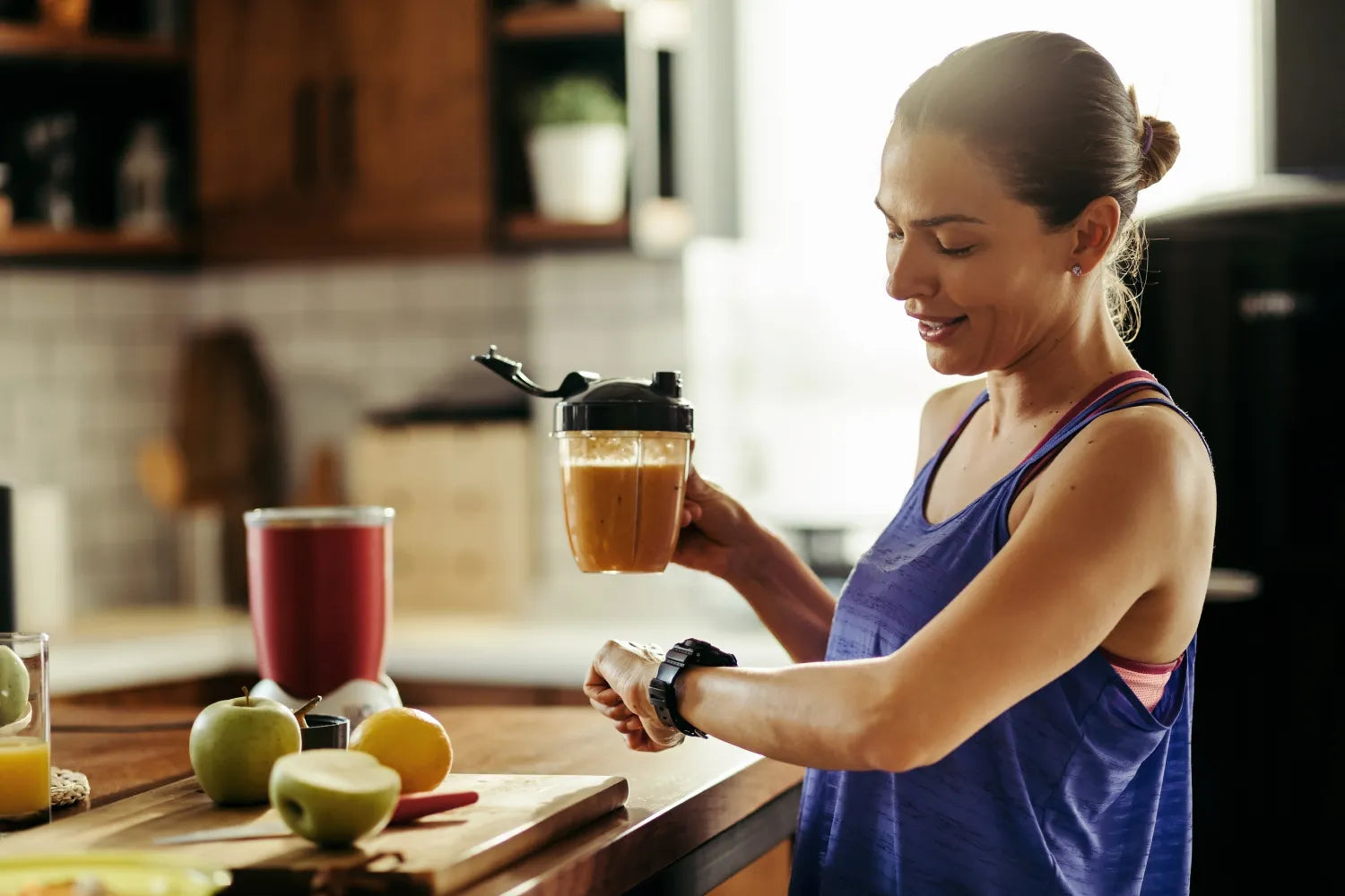 Woman in workout tank checking her watch while holding a blended morning drink in a kitchen with fruit on the counter