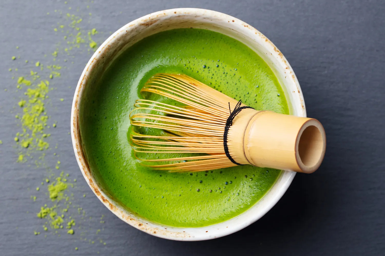 Bamboo whisk resting in a frothy bowl of bright green matcha on a dark slate background