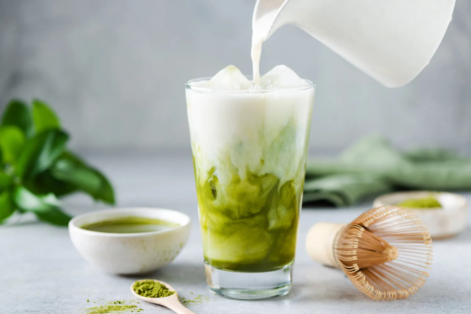 Iced matcha latte with milk being poured into a glass over ice, matcha powder and bamboo whisk on a light countertop