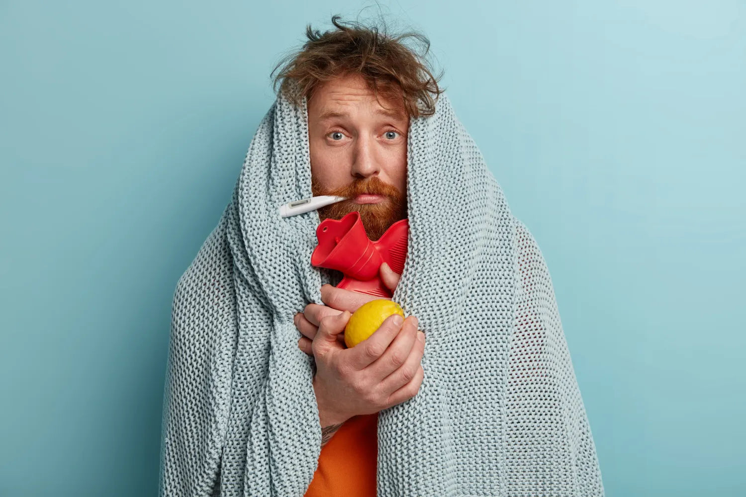 Bearded man wrapped in a light blue blanket holding a lemon and red hot water bottle with a thermometer in his mouth
