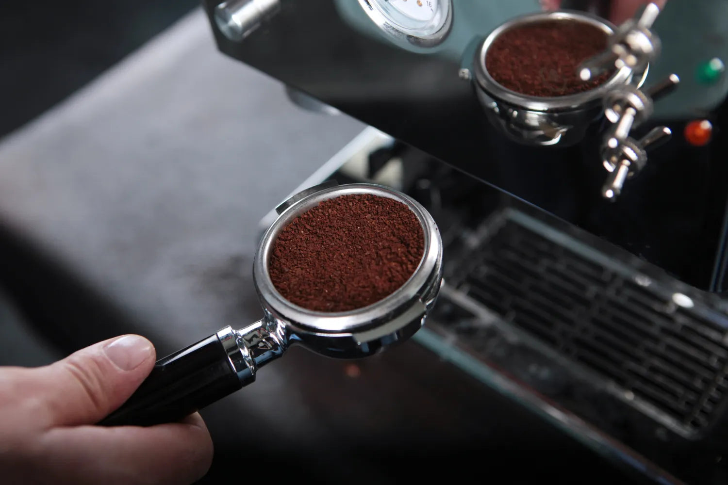 Hand holding an espresso portafilter filled with ground coffee beside an espresso machine