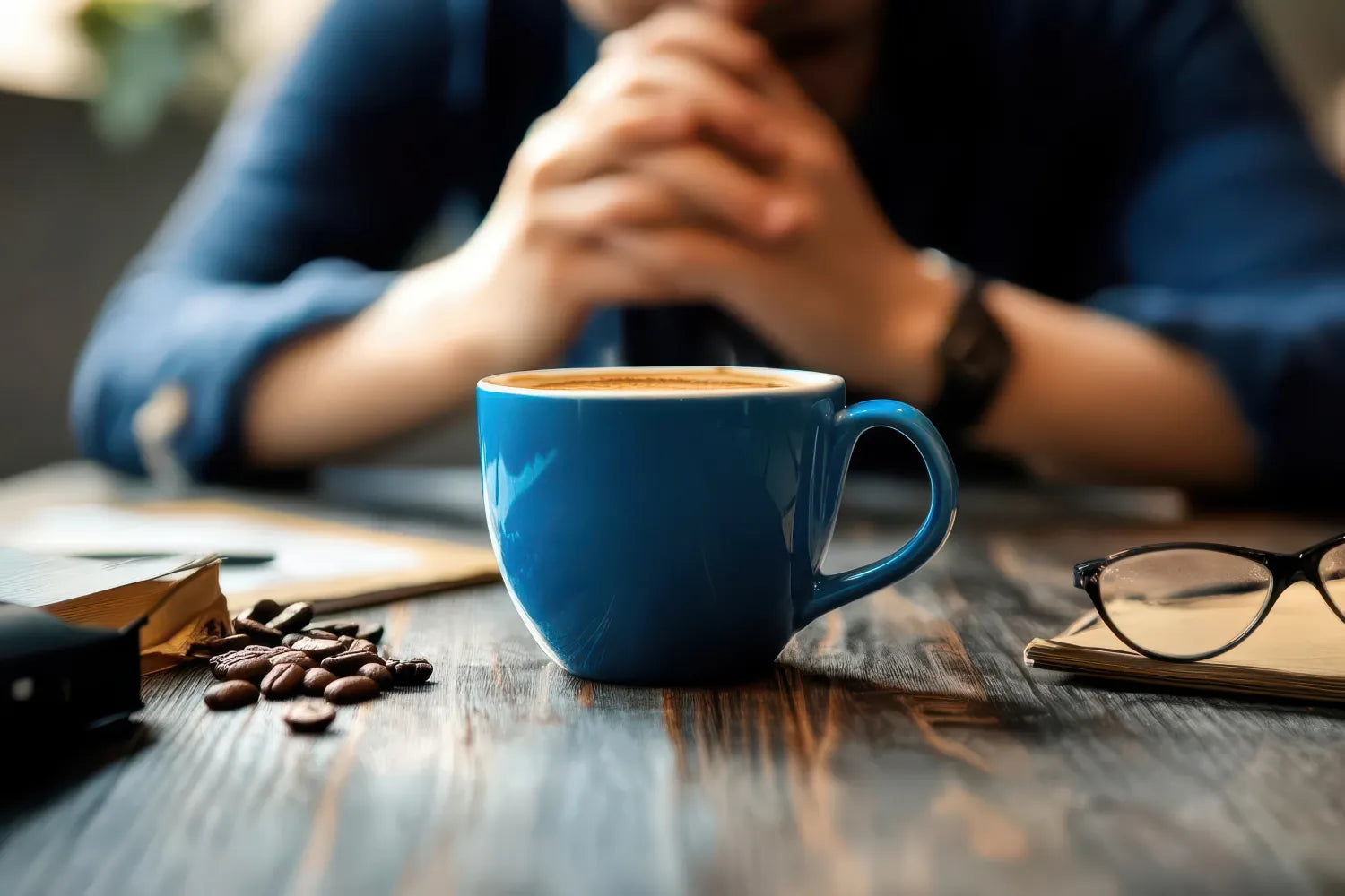 Blue coffee mug on a wooden table with coffee beans, a notebook and glasses, blurred person clasping hands in the background