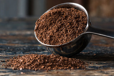 Close-up of a metal scoop filled with ground coffee showing medium fine brown grounds on a rustic wooden surface