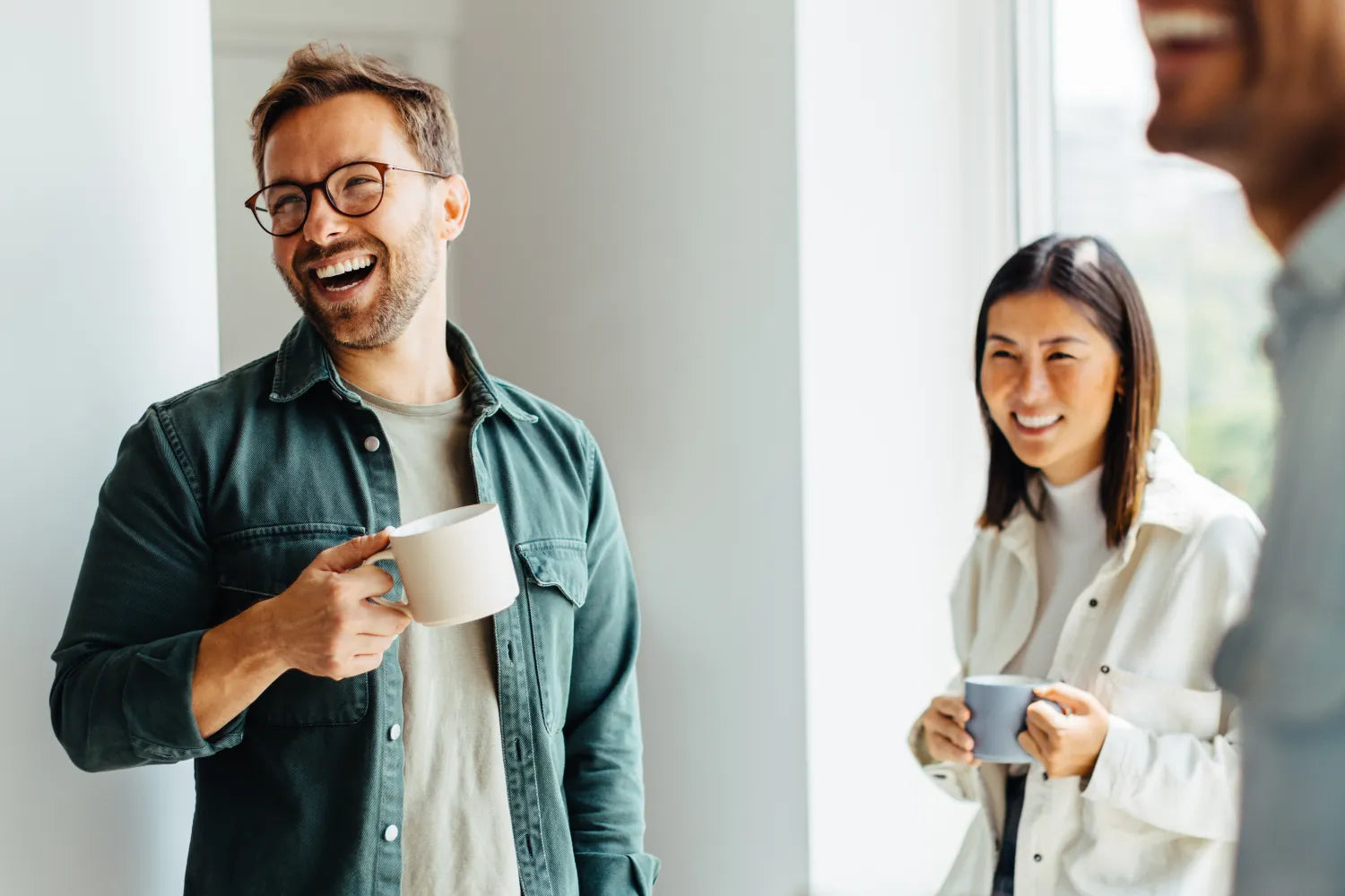 Two coworkers laughing while holding coffee mugs by a bright office window