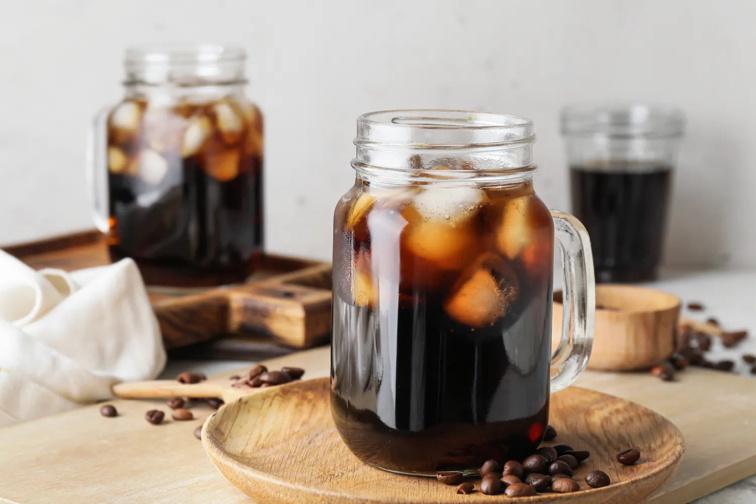 Mason jar of iced cold brew coffee on a wooden plate, with coffee beans and a second jar blurred in the background
