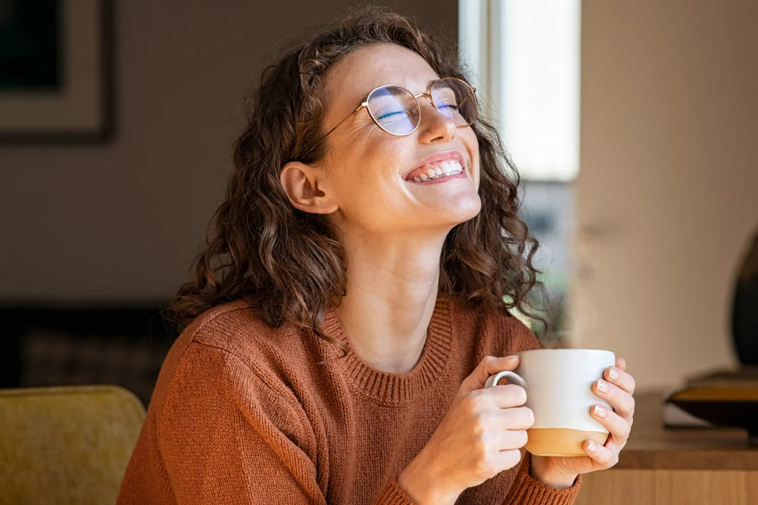 Woman in rust sweater smiling with eyes closed while holding a steaming mug of coffee at a kitchen table
