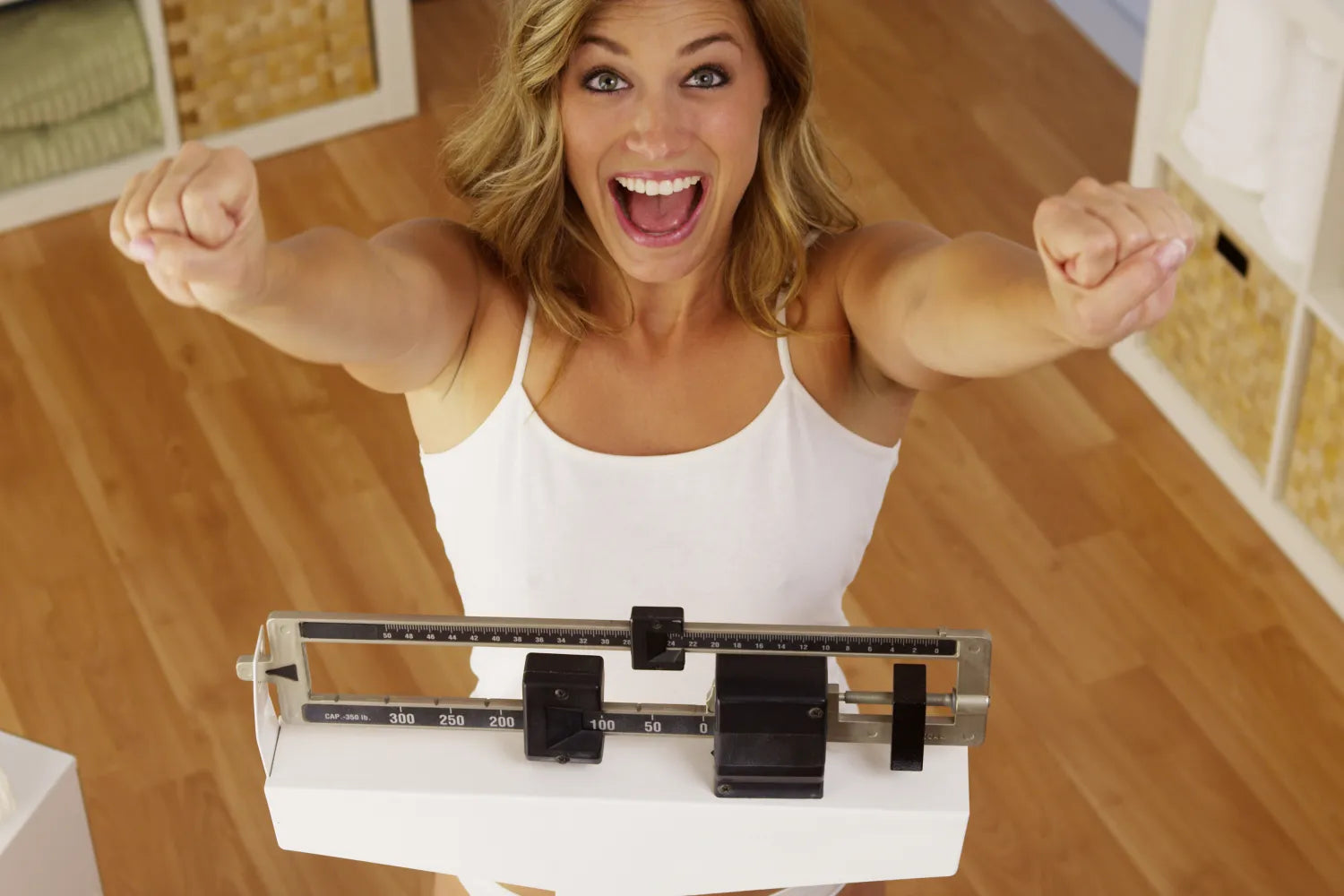 Smiling woman cheering on a bathroom scale after weighing herself, hardwood floor and storage baskets in background