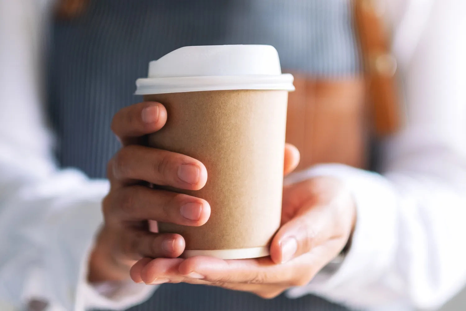 Person holding a brown paper to-go coffee cup with a white lid in both hands against a blurred background