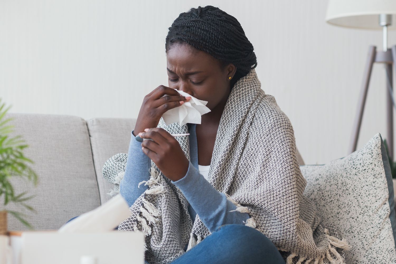Woman on a couch blowing her nose and checking a thermometer while wrapped in a blanket