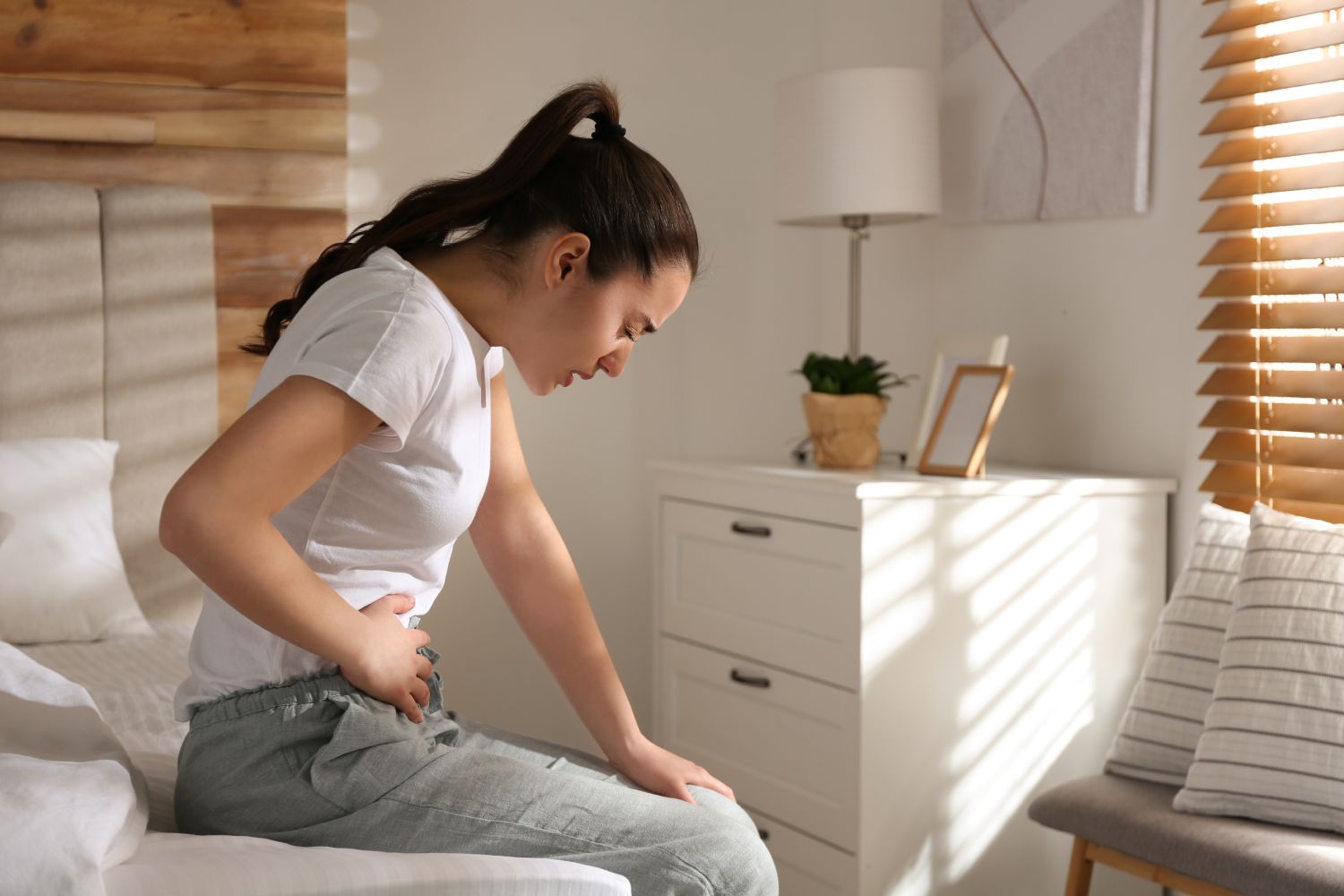 Young woman clutching her stomach and grimacing while sitting on a bed in a sunlit bedroom