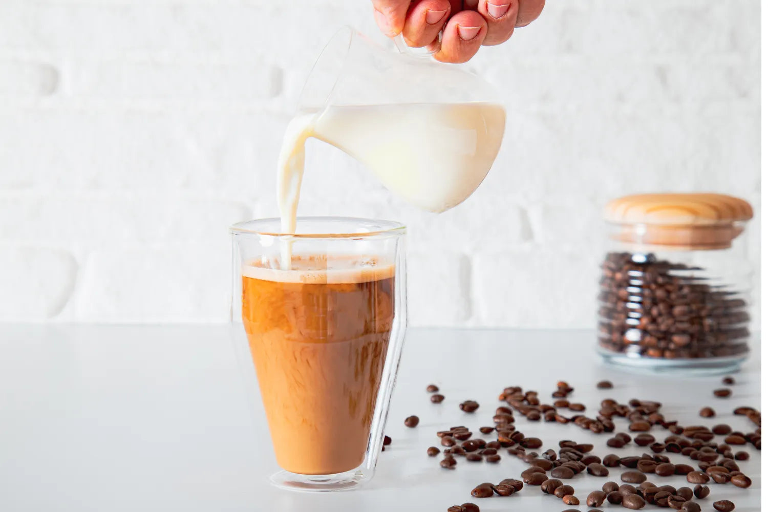 Hand pouring milk into brewed coffee in a double-walled glass with scattered coffee beans and a jar on a white countertop