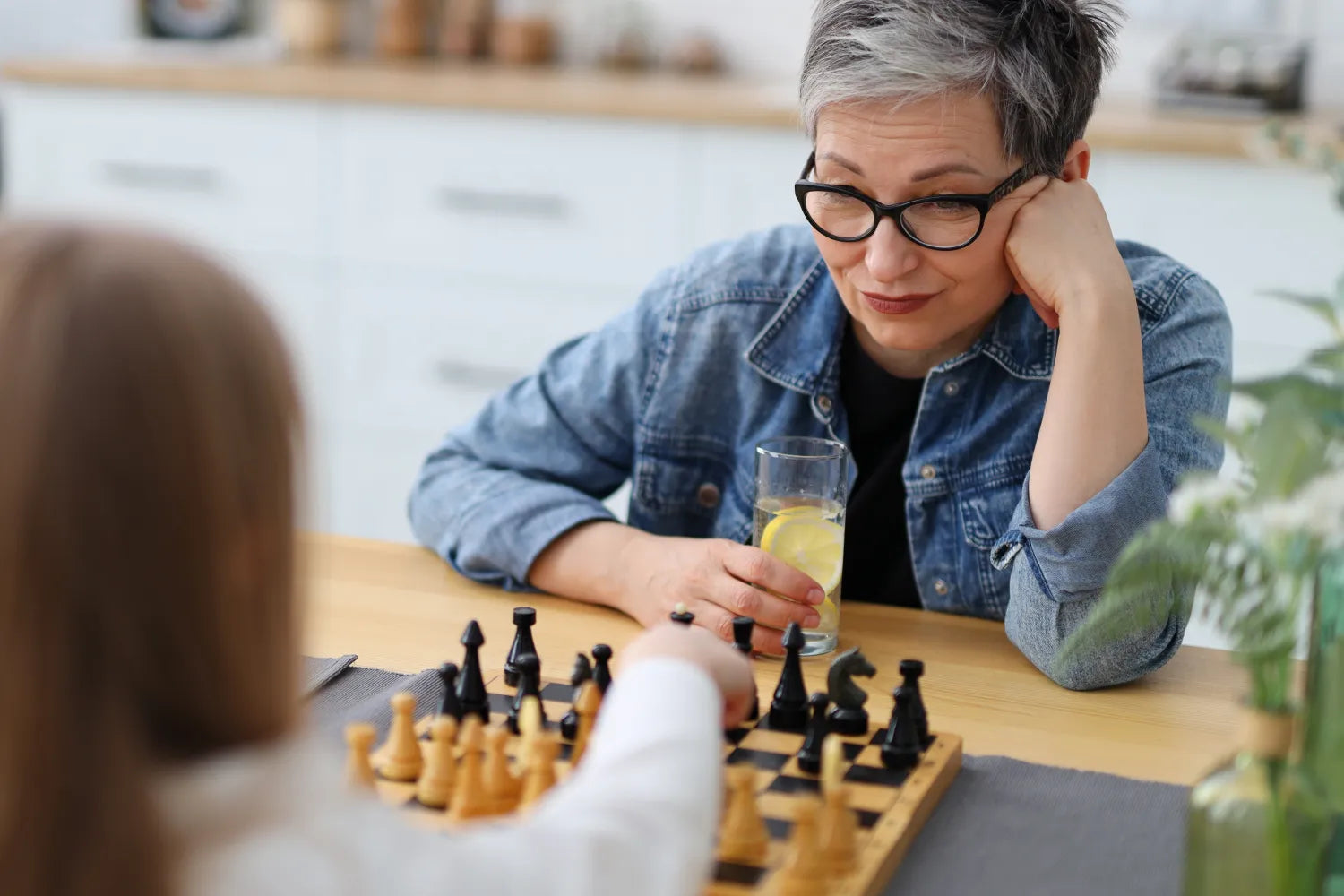 Older woman with glasses holding a glass of lemon water watching a child move a chess piece at a kitchen table