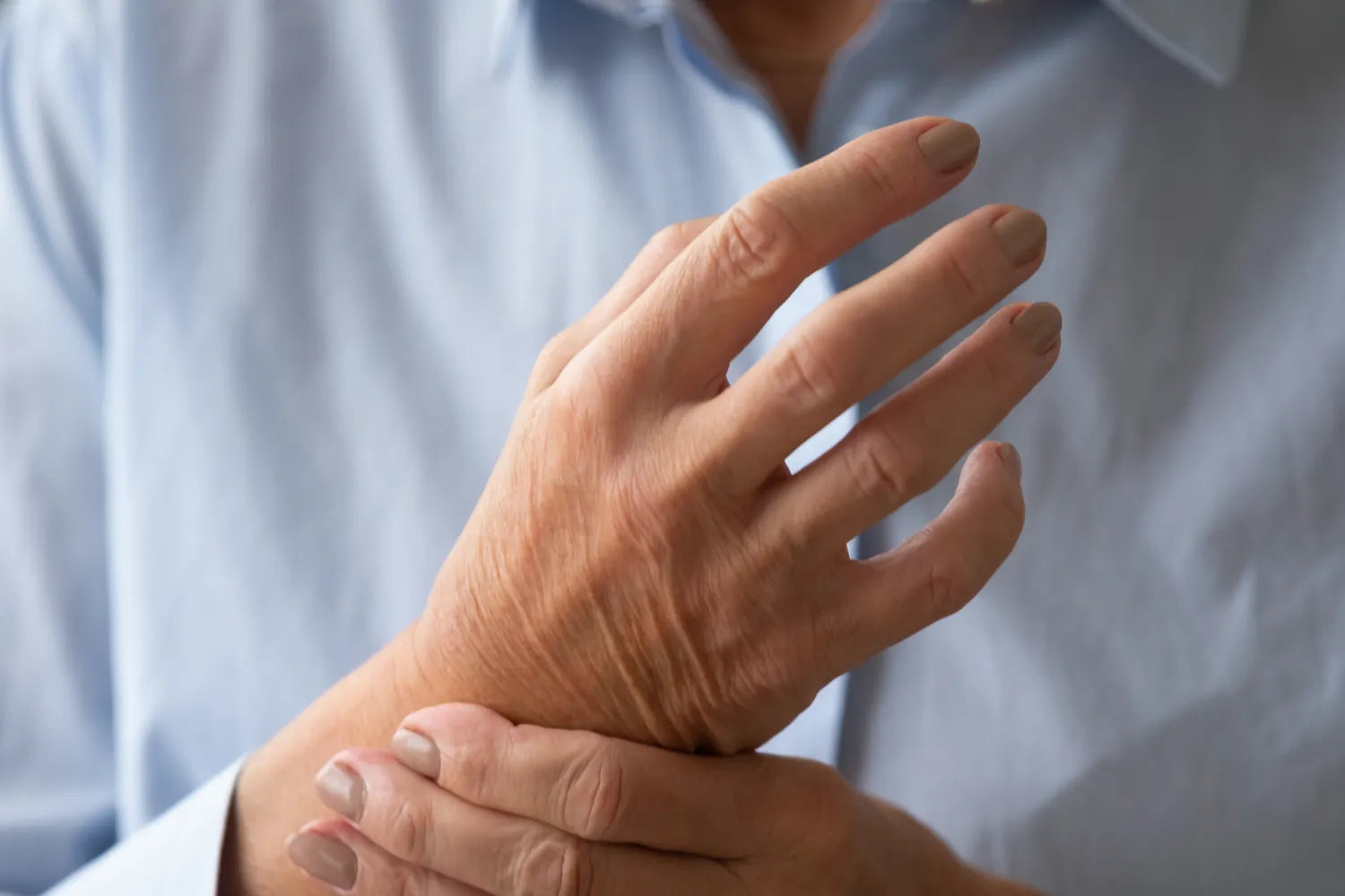 Close-up of an older adult holding their wrist in pain to illustrate arthritis, light blue shirt background