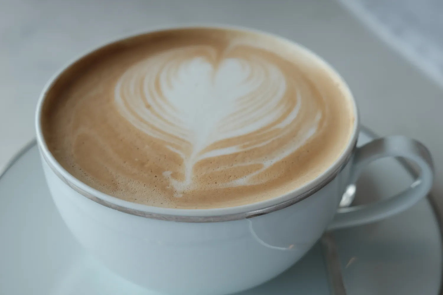 Close-up of cappuccino in white cup with heart-shaped latte art and saucer in soft-focus background
