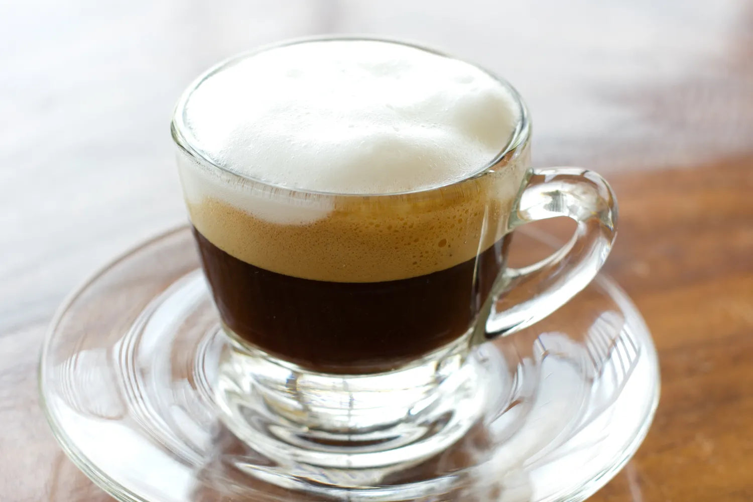 Glass cup of espresso topped with thick frothed milk on a clear saucer on a wooden table