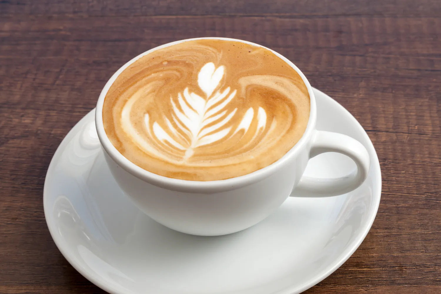 Top down view of a latte in a white cup showcasing rosette latte art with white saucer on a wooden table