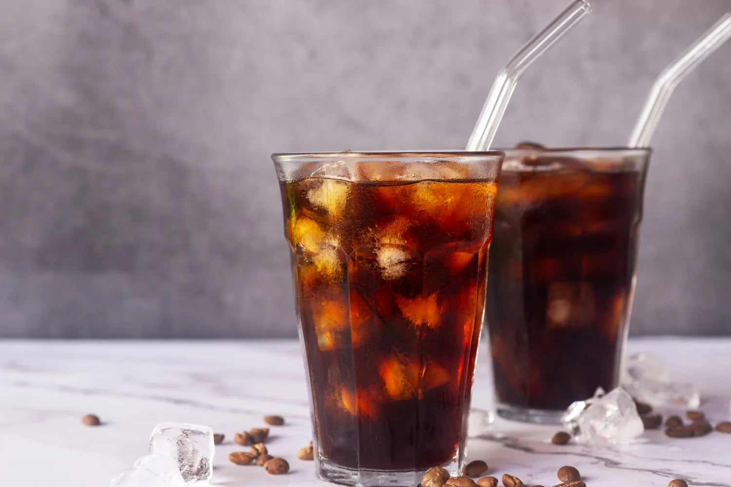 Two glasses of iced black coffee with ice and glass straws on a marble surface, coffee beans scattered around
