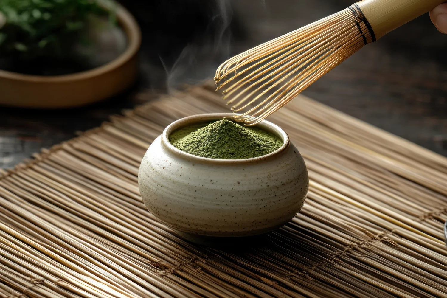 Close up of a bowl of vibrant green matcha powder with a bamboo whisk above on a bamboo mat