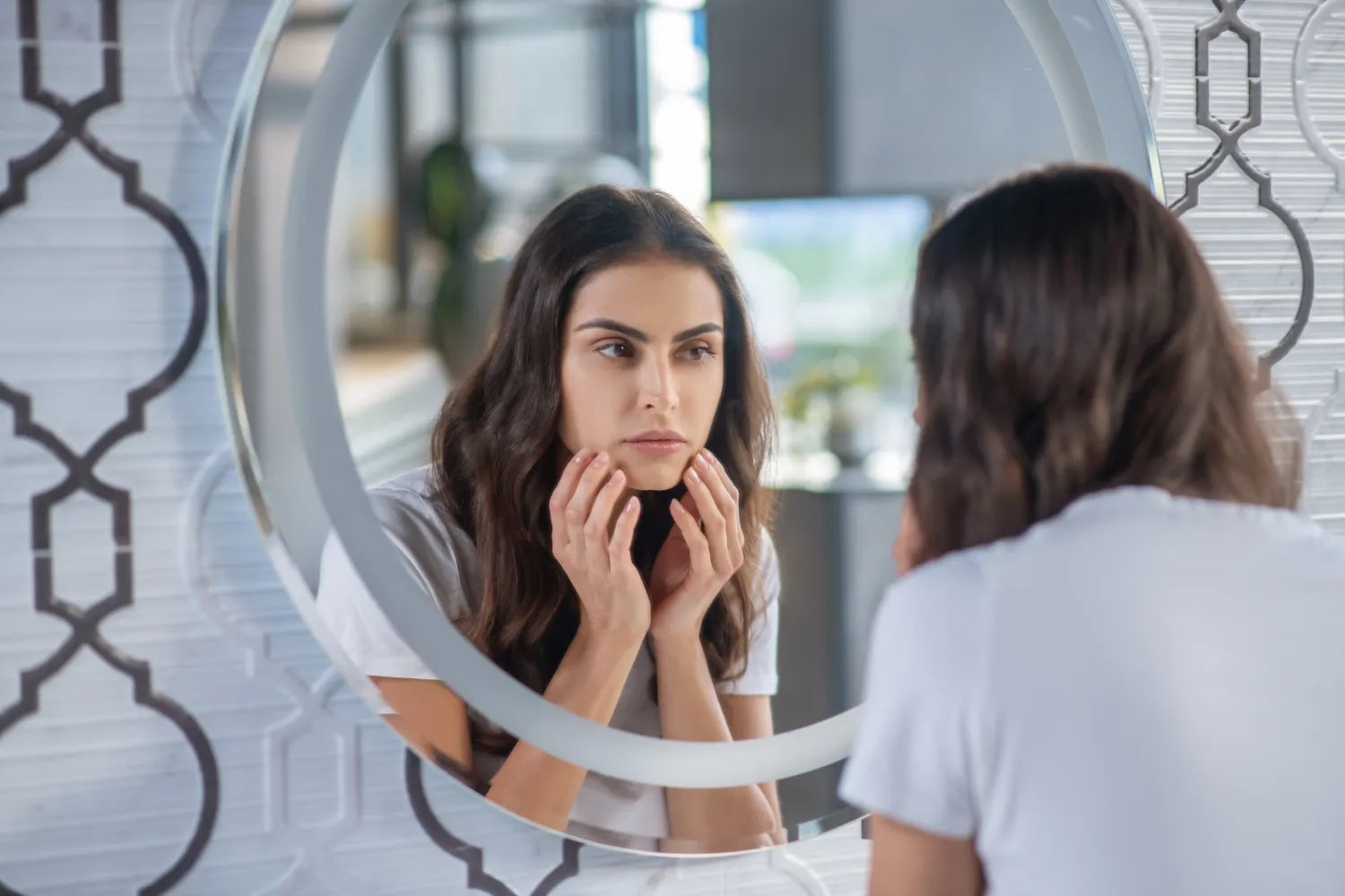 Woman examining her cheeks in a round mirror, touching her skin and looking concerned about loss of firmness