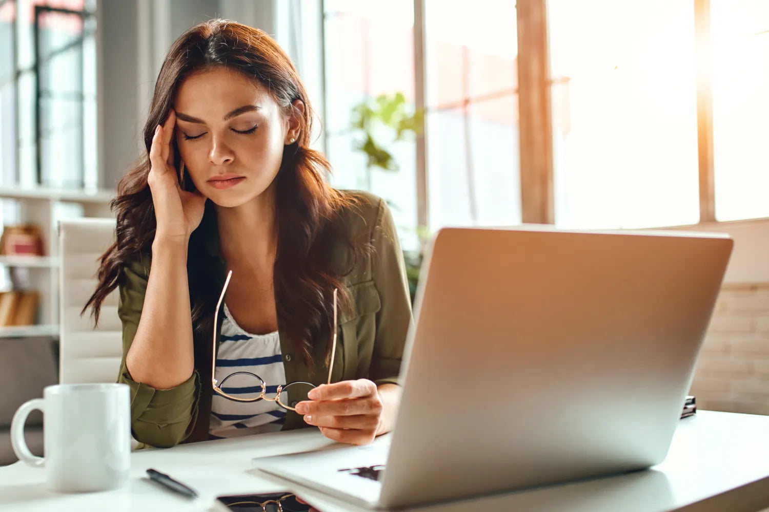 Woman rubbing her temple and holding glasses at a desk with laptop and coffee, looking stressed in a sunlit office