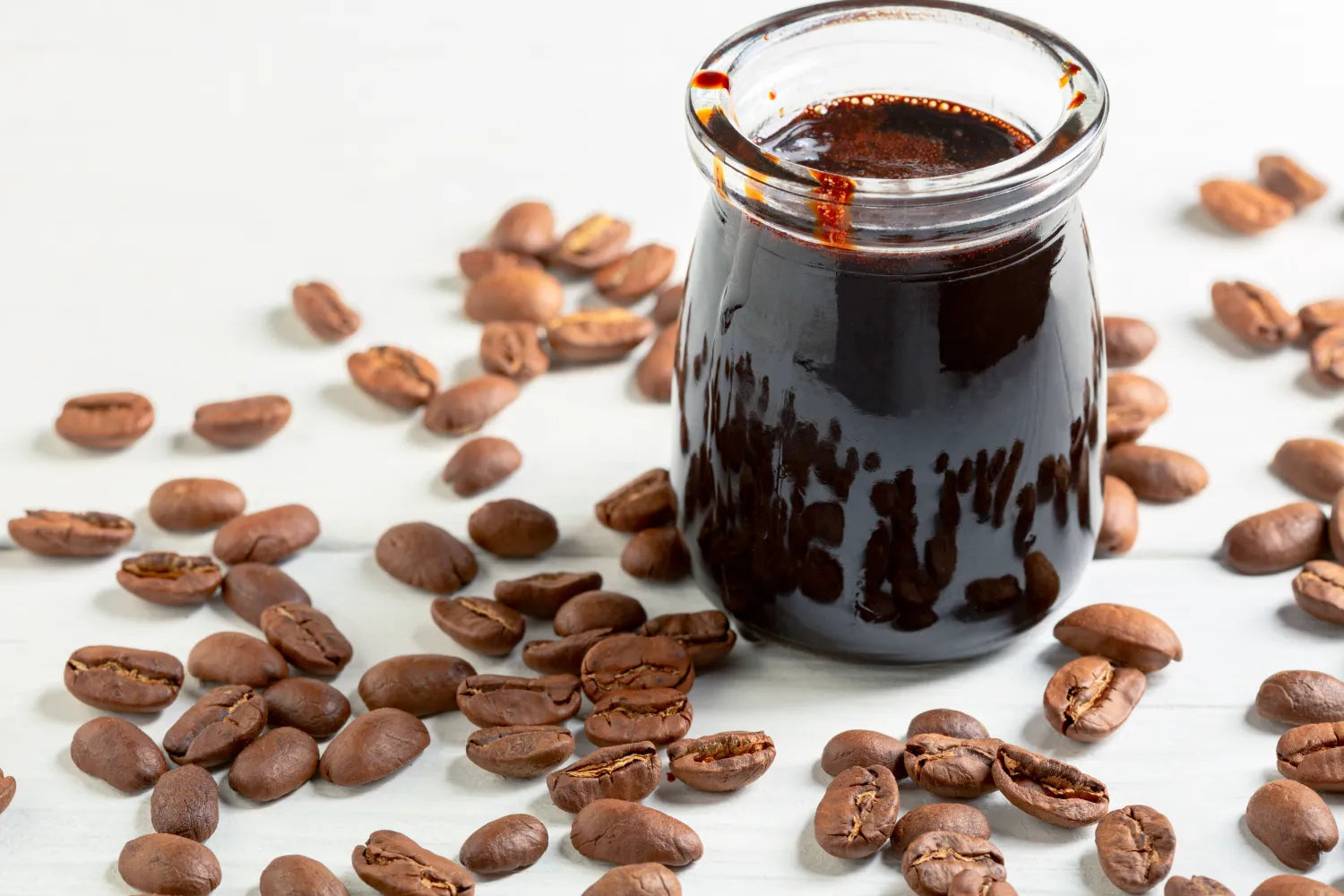 Close-up of glass jar of dark coffee extract showing thick glossy concentrate with roasted coffee beans on a white wooden surface